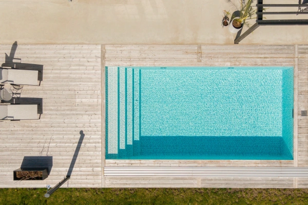 Pool med trädäck och sittplatser sedd ovanifrån i direkt solljus.