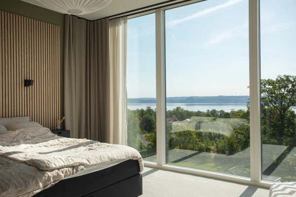 Modernt och ljust sovrum med en säng bäddad i linne, en sänggavel av träribbor och ett panoramafönster med vidsträckt utsikt över en sjö och trädtoppar.