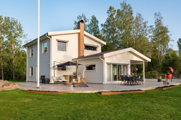 Vit tvåplansvilla med träfasad, stora fönsterpartier och rymlig uteplats med matplats och utekök.