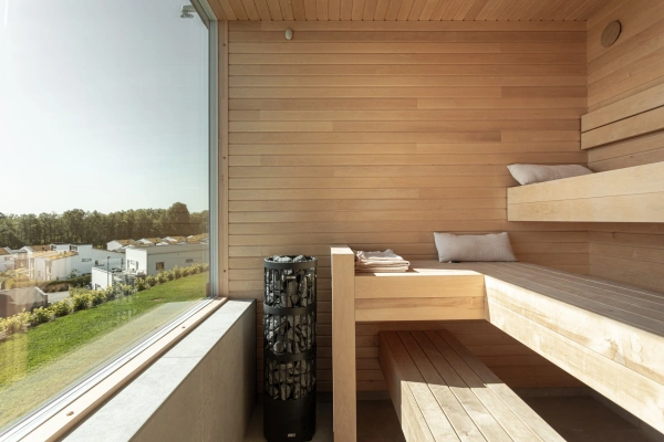 Modern bastu i ljust trä med lavar i etage och ett cylindriskt bastuaggregat. Ett stort fönster till vänster ger rikligt med ljus.