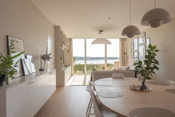 Ljus, öppen planlösning med en matgrupp i förgrunden och ett vardagsrum längre in. Ett stort fönsterparti i bakgrunden ger en vidsträckt utsikt över Vättern och grönskande natur.