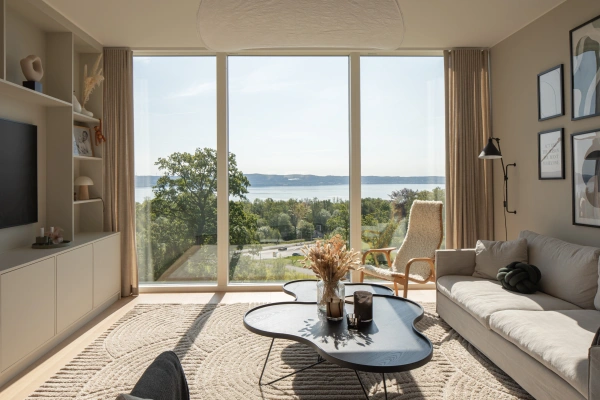 Ljust vardagsrum med en beige soffa, speciellt formade soffbord och en medieenhet, framför ett stort fönster med vidsträckt utsikt över Vättern, grönska och landskapet.