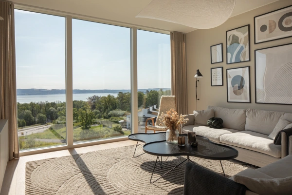 Ljust och modernt vardagsrum med en beige soffa, svarta soffbord och en tavelvägg framför ett stort panoramafönster med vidsträckt utsikt över vättern och grönska.