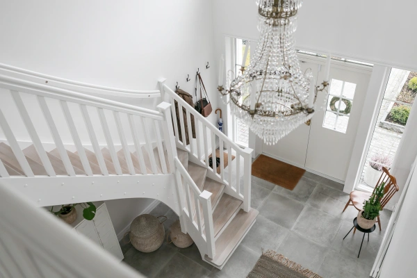 Rymlig entréhall sedd ovanifrån med en stor kristallkrona, vit trappa med ljusa trästeg, grått klinkergolv och ljus insläppande dubbeldörr