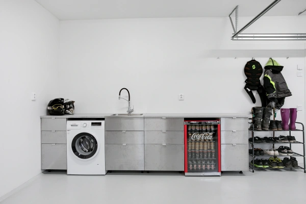 Arbetsbänk i rostfritt stål i ett garage med tvättmaskin, vask, rött Coca-Cola-kylskåp och skoställ.