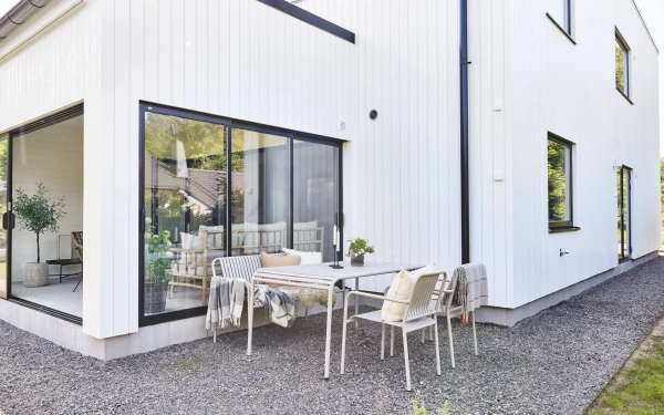 Vit träfasad med stora svarta fönsterpartier och uteplats med matgrupp intill huset.