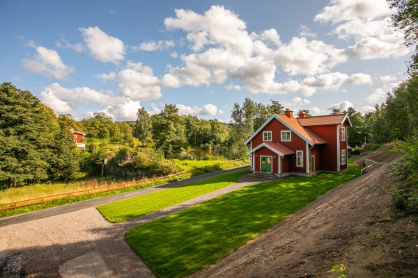 Rödmålat hus med vita knutar och grön dörr, beläget i ett lummigt landsbygdslandskap med vattendrag och gräsmatta.