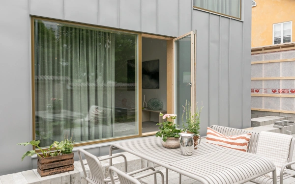 Uteplats med moderna ljusa utemöbler intill ett hus med grå plåtfasad och ett stort glasparti med öppen dörr