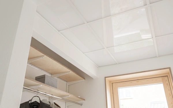 Närbild på hall med lysande pristak i rutor, hyllsystem i ljust trä och övre delen av en dörrkarm i trä
