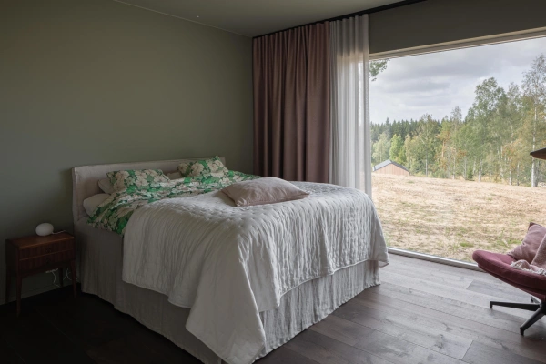 Harmoniskt sovrum med dovt gröna väggar, en bäddad dubbelsäng och ett stort panoramafönster mot natur och skog