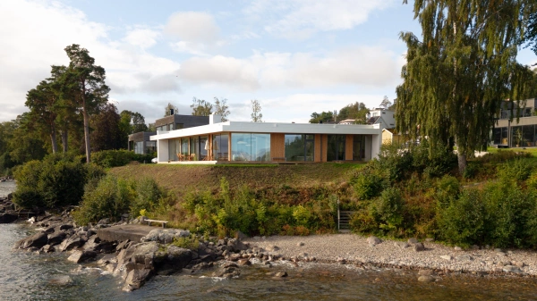 Flygbild av ett modernt enplanshus med stora glaspartier beläget på en sluttning vid en stenig strand.
