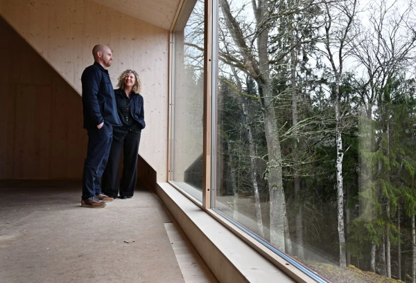 Två personer står i ett träklätt rum framför ett stort fönster med utsikt över skogen.