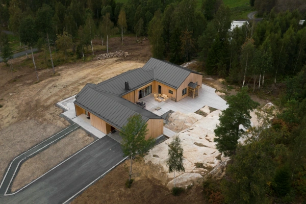 Flygbild över ett modernt trähus med mörkt plåttak, byggt intill en berghäll och omgivet av skog.