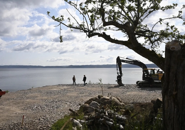 Flygbild över ett modernt trähus med mörkt plåttak, byggt intill en berghäll och omgivet av skog.